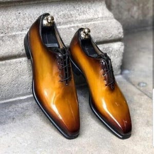 May include: A pair of brown and tan leather dress shoes with a glossy finish. The shoes have a classic Oxford design with a pointed toe, black laces, and a black sole. Shoe trees are inserted in the shoes.