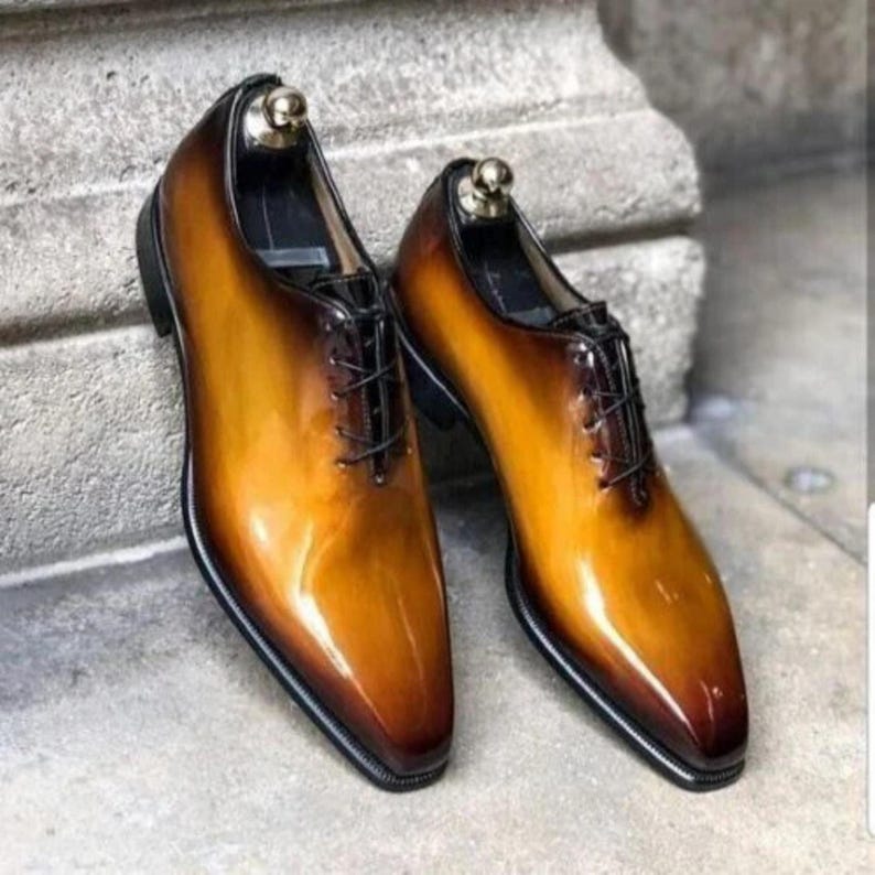 May include: A pair of brown and tan leather dress shoes with a glossy finish. The shoes have a classic Oxford design with a pointed toe, black laces, and a black sole. Shoe trees are inserted in the shoes.