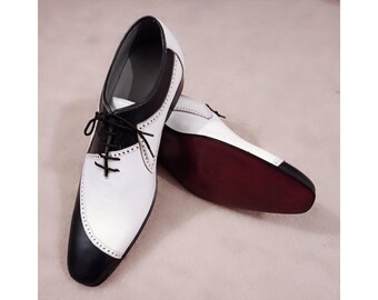 May include: A pair of two-tone spectator shoes featuring a black and white leather design with decorative brogue detailing along the seams. The shoes have black laces, a rounded toe, and a dark reddish-brown sole visible on the second shoe.