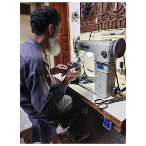 May include: A craftsman using a light grey sewing machine. The machine has a large wheel and a black belt. The text "Bespoke Handmade" is visible. The craftsman is wearing a grey shirt and has a white beard.