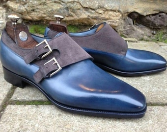 May include: A pair of blue leather double monk strap shoes featuring dark grey suede overlays on the upper, silver-toned metal buckles, and dark wooden shoe trees inserted inside each shoe, set against a textured stone background.