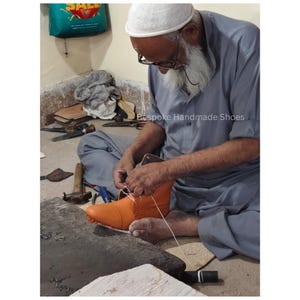 May include: An artisan meticulously hand-sewing an orange leather boot. The craftsman, wearing glasses and a white cap, is focused on the stitching process. The image conveys the creation of bespoke, handmade footwear. The text "Bespoke Handmade Shoes" is visible.