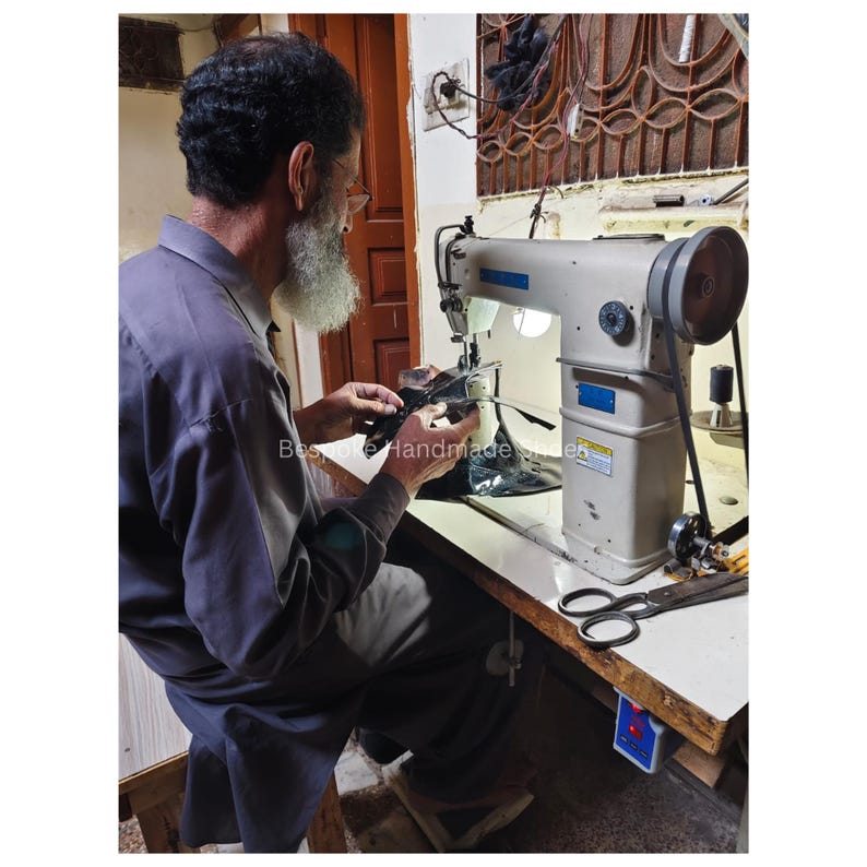 May include: An artisan working at a sewing machine. The machine is light gray with a large wheel and a black belt. The artisan is wearing a gray shirt and has a white beard. The text "Bespoke Handmade shoes" is visible.