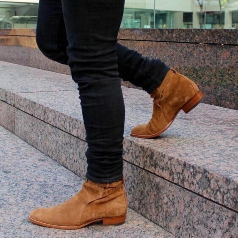 May include: A person wearing brown suede ankle boots with a low heel and a rounded toe. The boots are paired with black denim jeans. The person is walking up a set of stone steps.