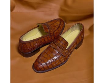 May include: A pair of brown loafers featuring a glossy crocodile-embossed texture. These slip-on shoes have a classic penny loafer strap across the vamp, a rounded toe, a low stacked heel, and a light yellow interior lining with visible stitching.