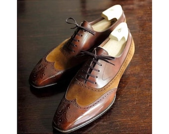 May include: A pair of brown leather dress shoes with a wingtip design. The shoes feature a two-tone color scheme with a lighter brown suede panel and a darker, polished leather. The shoes have laces and are displayed with wooden shoe trees.