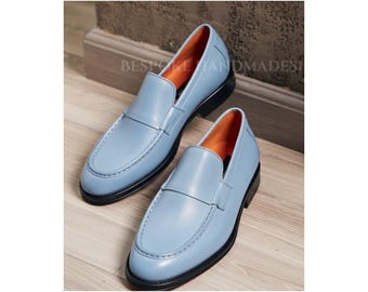 May include: A pair of light blue leather loafers featuring a smooth finish, a rounded toe, and a strap across the vamp. The interior lining is bright orange, and the shoes are set on a black sole. The text BESPOKE HANDMADES is visible in the background.
