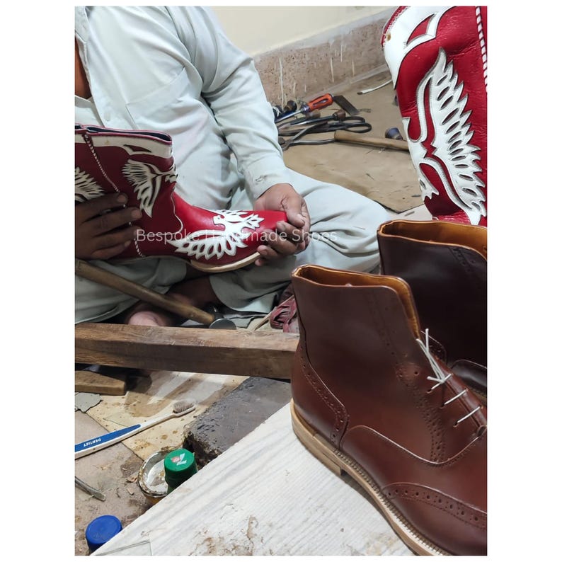 May include: A craftsman working on leather boots. One red boot with white decorative cutouts is held in the hands. Another brown boot is in the foreground. The text "Bespoke Handmade Shoes" is visible on the red boot.