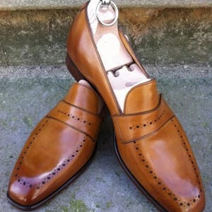 May include: A pair of brown leather loafers with decorative perforations and a polished finish. The shoes have a classic design with a rounded toe and a slip-on style. Shoe trees are inserted inside the shoes to maintain their shape.