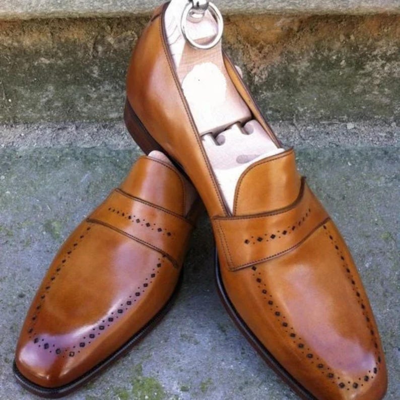 May include: A pair of brown leather loafers with decorative perforations and a polished finish. The shoes have a classic design with a rounded toe and a slip-on style. Shoe trees are inserted inside the shoes to maintain their shape.