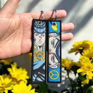 May include: Two black and blue keychains with a printed image of a character from the anime JoJo's Bizarre Adventure. The character has blue hair and is wearing a blue and gold outfit. The keychains are hanging from silver key rings.