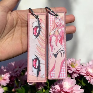 May include: Two pink anime-style keychains. One keychain features a close-up of a woman's eyes with red and black pupils. The other keychain features a woman in a pink bikini with long pink hair. Both keychains have a white background with pink polka dots.