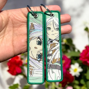 May include: Two green fabric keychains with white anime character illustrations. The characters have long white hair and are wearing white clothing. The keychains are attached to silver key rings.