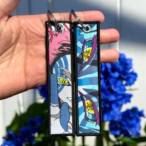 May include: Two black and white keychains with a blue and white striped design. One keychain features a character with pink hair and a blue and white face mask. The other keychain features a blue and white eye with a yellow pupil. Both keychains have the Japanese word "鬼滅" written on them.