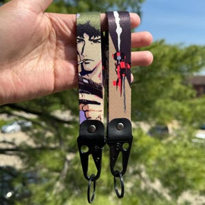 May include: Two wrist lanyards with black hardware. One lanyard features an anime character with a cigarette, the other has a red and black graphic. Both lanyards are held up against a green background.