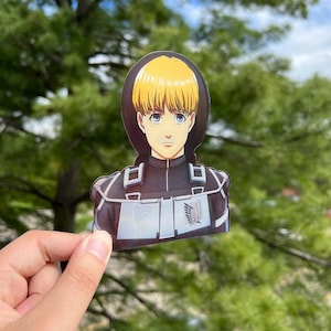 May include: A sticker featuring a cartoon character with blonde hair, wearing a black and white uniform with a symbol on the chest. The character has a serious expression.