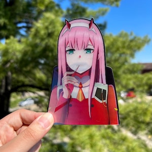 May include: A pink-haired anime character with green eyes and a red and white outfit is holding a lollipop in her mouth. The character is depicted in a sticker format.