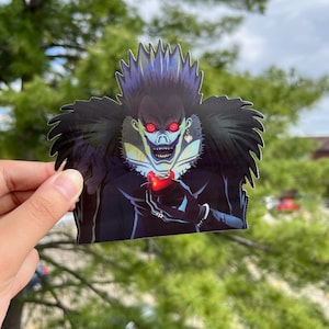 May include: A sticker featuring a black and white illustration of a character with red eyes and a menacing grin, holding a red apple. The character is wearing a black coat with a large collar and a heart-shaped earring.