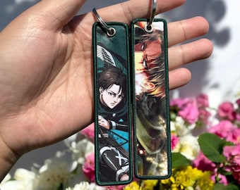 May include: Two green fabric keychains with anime characters printed on them. The keychain on the left features a character with brown hair and green eyes. The keychain on the right features a character with blonde hair and blue eyes.
