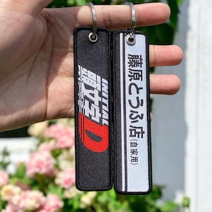 May include: Two black and white fabric keychains with silver key rings. The keychain on the left has a red and white logo that says "Initial D" in Japanese. The keychain on the right has white text that says "藤原とうふ店 (豆腐)" in Japanese.