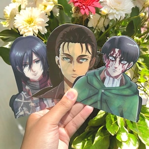 May include: Three anime character cutouts are held in front of a floral background. The characters are Mikasa, Eren, and Levi. Eren has brown hair and blue eyes. Levi has blood on his face. Mikasa has black hair.