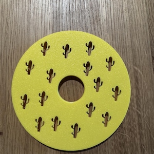 Op de afbeelding: Een gele onderzetter met een centraal gat en kleine cactusuitsparingen. De onderzetter is rond en heeft een getextureerd oppervlak. De cactusuitsparingen zijn gelijkmatig verdeeld.