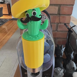 May include: A novelty bottle stopper featuring a green cactus with a black moustache and a yellow sombrero. The cactus has arms and a smiling face. The stopper is yellow and green.