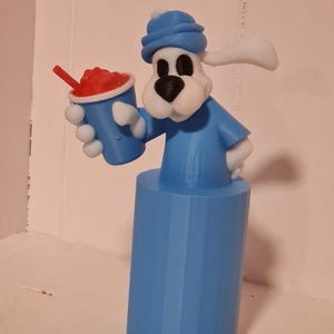 Puede incluir: Figura impresa en 3D de un perro de dibujos animados con gorro y camisa azules, sosteniendo una taza azul con un granizado rojo y una pajita. El perro es blanco con ojos y nariz negros, y la base es un cilindro azul sólido.