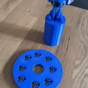 Peut inclure: Une figurine bleue de Stitch de Lilo & Stitch, posée sur un cylindre bleu. Un dessous de verre bleu avec des découpes en forme de feuilles est également présent. Les articles sont en plastique et sur une surface en bois.