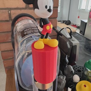 May include: A red and yellow Mickey Mouse figure stands atop a red cylindrical object. The figure has black ears, a white face, and wears red shorts, yellow shoes, and a black shirt. The object is attached to a clear appliance.