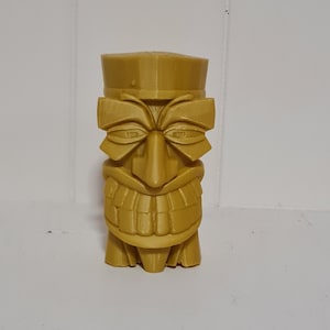 Puede incluir: Una estatua Tiki dorada con un sombrero cuadrado, ojos grandes y una amplia sonrisa. La estatua tiene una apariencia tallada detallada y se encuentra sobre una superficie blanca. La figura Tiki es una representación de la cultura polinesia.