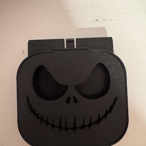 Peut inclure: Objet décoratif noir de forme carrée avec un visage de Jack Skellington sculpté, tiré de L'Étrange Noël de Monsieur Jack. Le design comprend de grandes orbites et un large sourire. L'objet a une surface texturée et un dessus rectangulaire.
