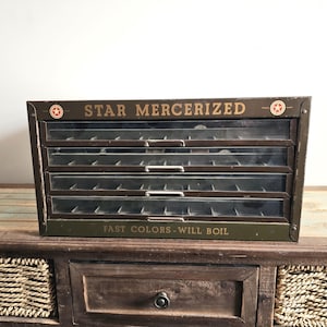 May include: Vintage metal cabinet with four glass-fronted drawers. The cabinet is olive green with gold lettering that reads "STAR MERCERIZED" and "FAST COLORS - WILL BOIL". The cabinet is sitting on a wooden table.