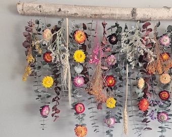 May include: A dried flower wall hanging with a birch branch at the top. The flowers are in shades of pink, yellow, orange, and red. The flowers are hanging from eucalyptus branches.