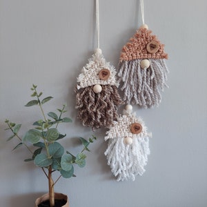May include: Three macrame gnome ornaments hanging by white strings. Each gnome features a triangular woven hat with a wooden heart-shaped button, a round wooden bead nose, and a long, textured fringe beard in cream, brown, gray, or white.
