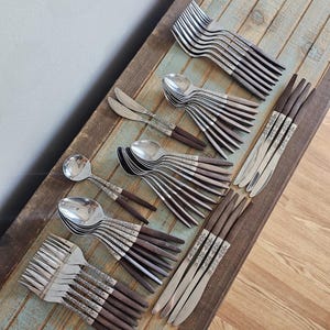 May include: A collection of vintage silverware, including forks, spoons, and knives, arranged on a rustic wooden surface. The silverware features ornate silver detailing and dark brown wooden handles. The arrangement suggests a formal dining setting.