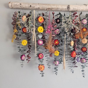 May include: A dried flower wall hanging with a birch branch at the top. The hanging features a variety of dried flowers in shades of pink, yellow, orange, and red. The flowers are arranged in a cascading pattern, creating a beautiful and natural look.