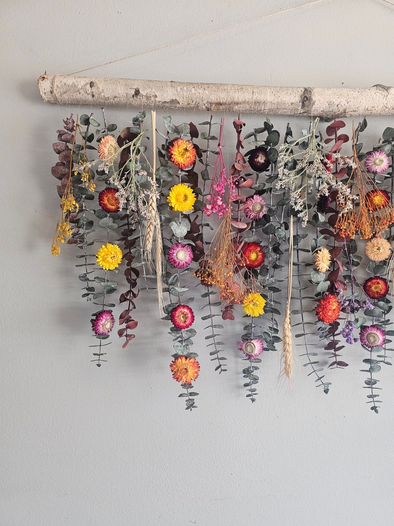 May include: A dried flower wall hanging with a birch branch at the top. The hanging features a variety of dried flowers in shades of pink, yellow, orange, and red. The flowers are arranged in a cascading pattern, creating a beautiful and natural look.