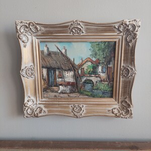 May include: A framed oil painting depicting a quaint village scene with thatched roof houses, a small bridge, and a stream. The painting is in a gold-colored frame with ornate details.