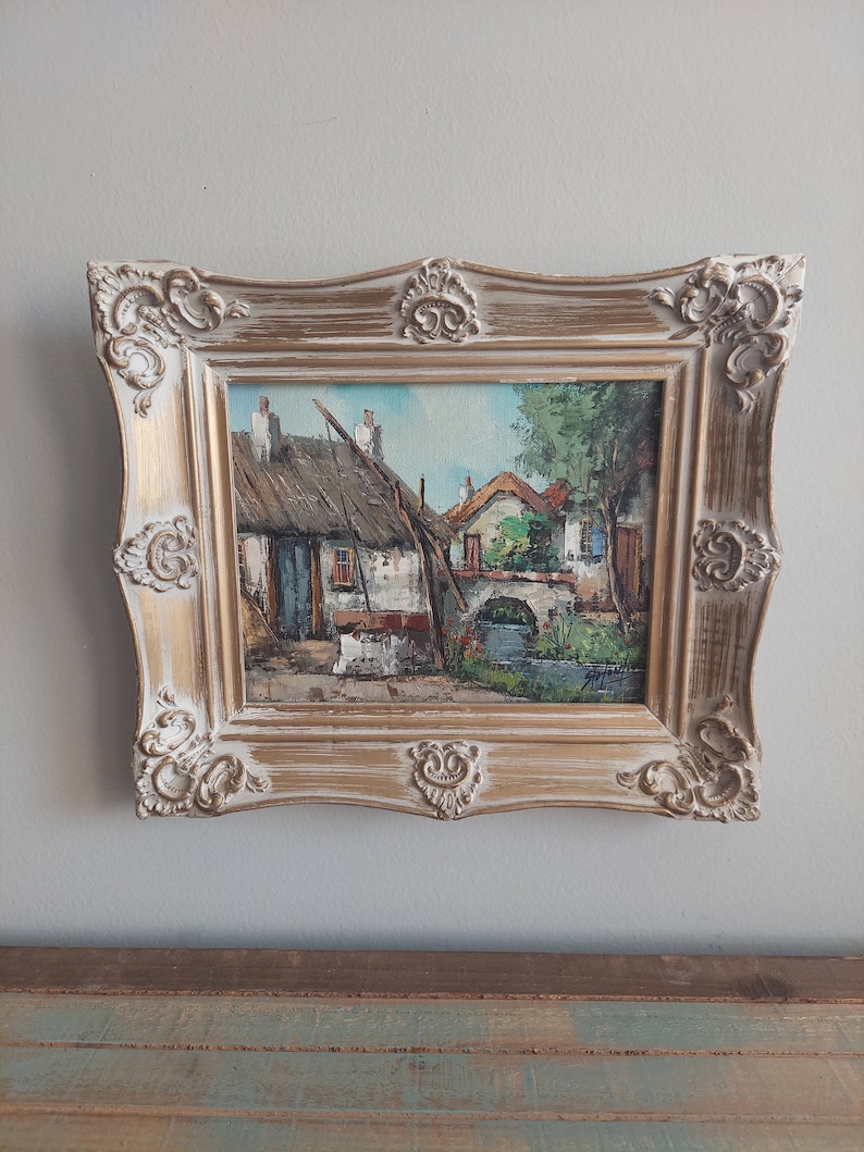 May include: A framed oil painting depicting a quaint village scene with thatched roof houses, a small bridge, and a stream. The painting is in a gold-colored frame with ornate details.