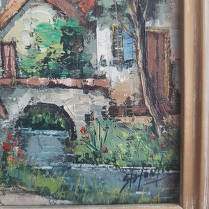 May include: A painting of a small village scene with a stone bridge over a stream, a white house with a brown door, and a green tree. The painting is framed in a light brown wood frame.