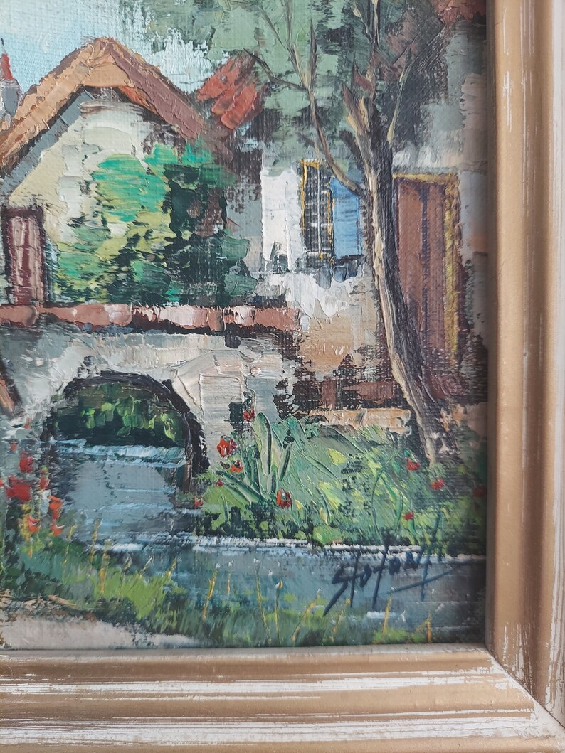 May include: A painting of a small village scene with a stone bridge over a stream, a white house with a brown door, and a green tree. The painting is framed in a light brown wood frame.