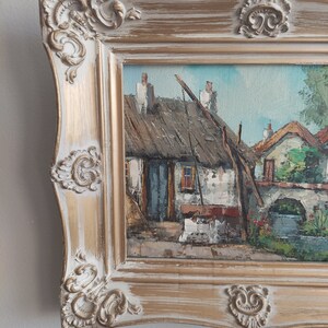 May include: A framed oil painting depicting a quaint village scene with a thatched roof cottage, whitewashed walls, and a stone bridge. The painting is set in a gold-toned frame with ornate details.