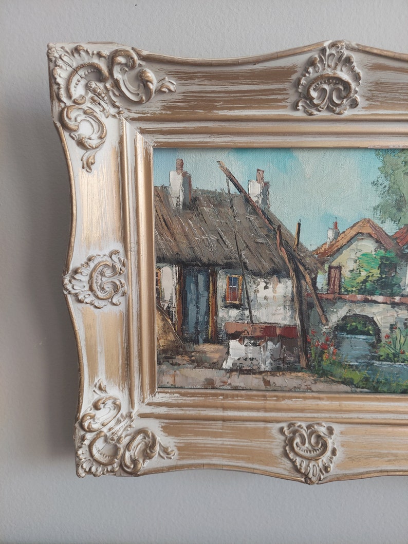 May include: A framed oil painting depicting a quaint village scene with a thatched roof cottage, whitewashed walls, and a stone bridge. The painting is set in a gold-toned frame with ornate details.