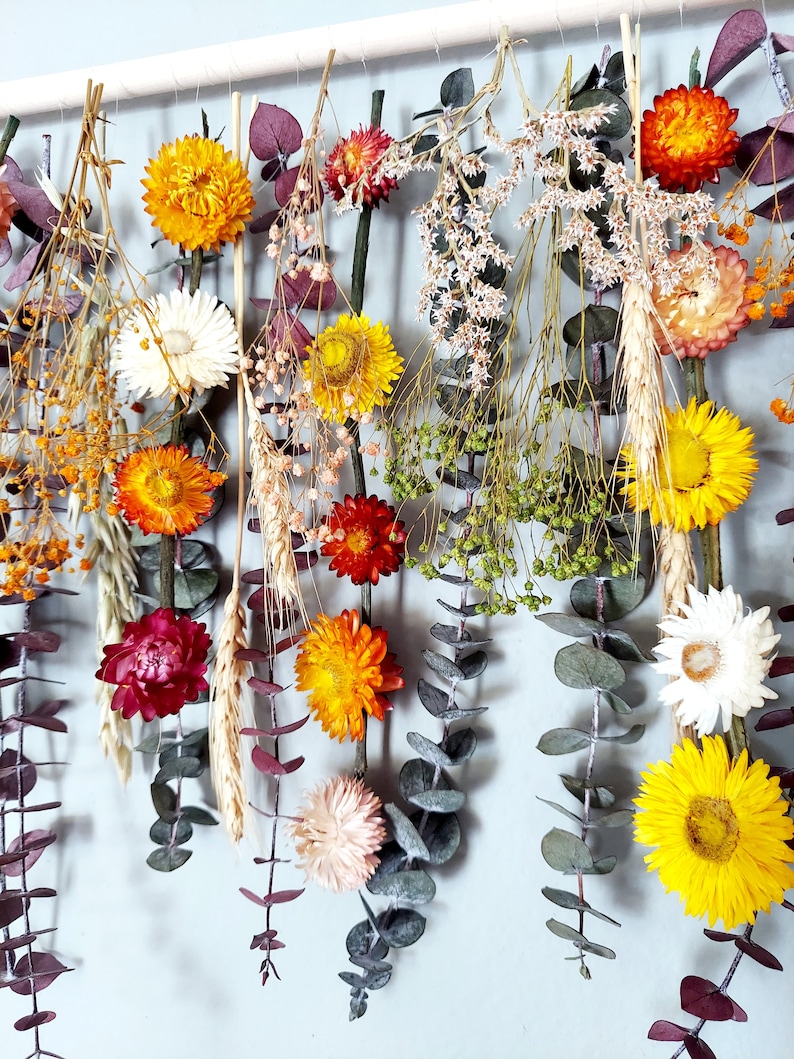 May include: A wall hanging made with dried flowers and greenery. The flowers are in shades of yellow, orange, red, and white. The greenery is a variety of eucalyptus leaves.