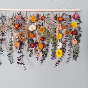 May include: A dried flower garland with a variety of colors, including yellow, orange, red, and white. The garland is hanging from a wooden dowel rod and is made with eucalyptus leaves and dried flowers.