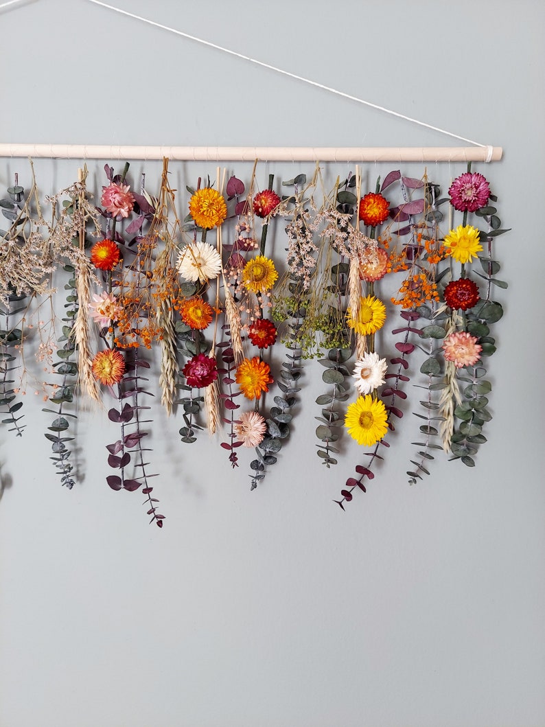 May include: A dried flower garland with a variety of colors, including yellow, orange, red, and white. The garland is hanging from a wooden dowel rod and is made with eucalyptus leaves and dried flowers.