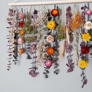 May include: A wall hanging made of dried flowers and greenery. The flowers are in shades of yellow, orange, red, and pink. The greenery is a mix of eucalyptus and other types of leaves.