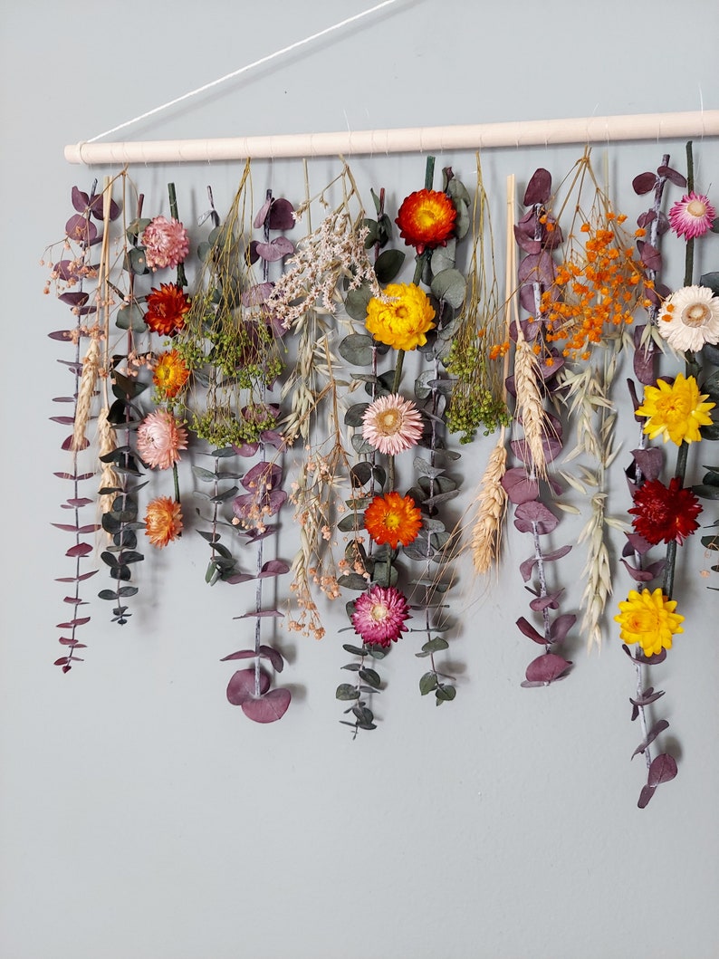May include: A wall hanging made of dried flowers and greenery. The flowers are in shades of yellow, orange, red, and pink. The greenery is a mix of eucalyptus and other types of leaves.
