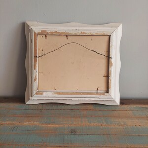 May include: White wooden picture frame with a scalloped edge. The frame is slightly distressed and has a wire hanger on the back.